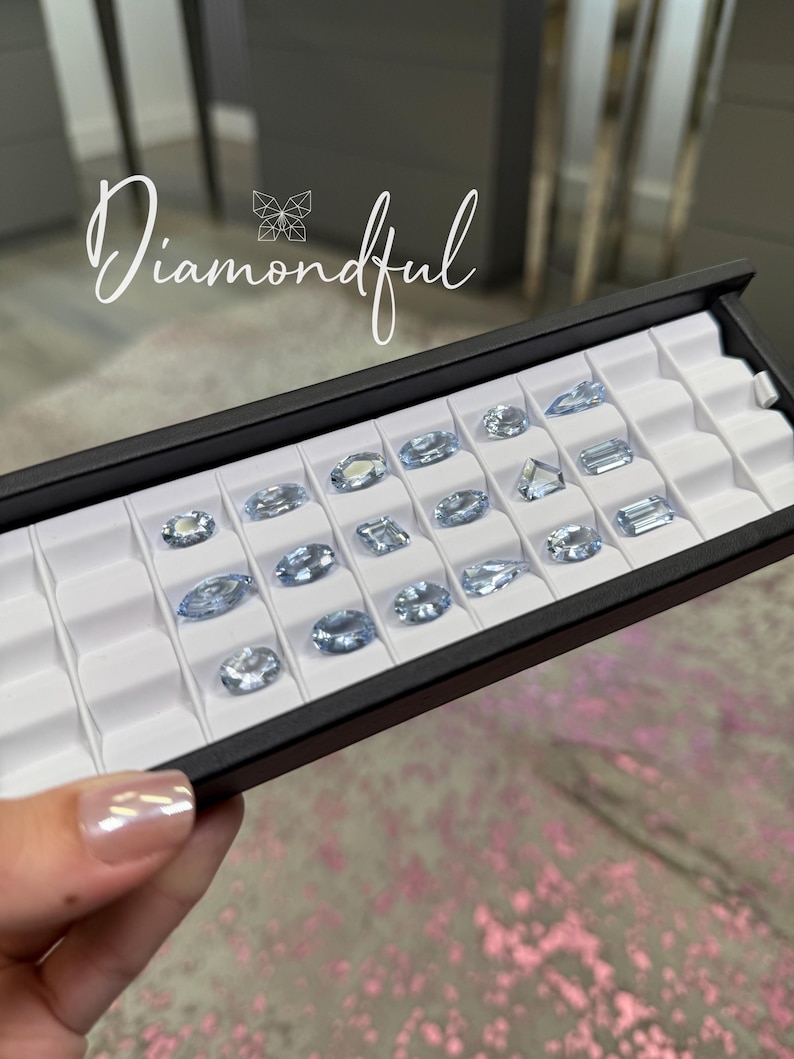 Op de afbeelding: Een zwarte tray gevuld met diverse lichtblauwe edelstenen in verschillende vormen, waaronder ovaal, peer en rechthoekig. De binnenkant van de tray is wit. Het woord "Diamondful" is in cursief geschreven. De tray wordt door een persoon vastgehouden.
