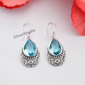 May include: A pair of silver drop earrings featuring a light blue, teardrop-shaped gemstone. The earrings have an intricate, filigree pattern around the stone. The background is white with a red flower.