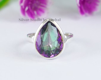May include: A silver ring with a large, pear-shaped gemstone. The gemstone displays a mix of colors, including purple, green, and blue. The ring is set against a white background with blurred purple flowers and green stems. The text "Silver Studio by Snehal" is visible.