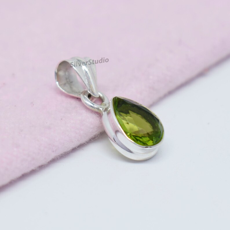 May include: A silver pendant with a pear-shaped green gemstone. The pendant is on a pink surface.