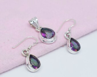 May include: A jewelry set featuring a pendant and a pair of earrings. Each piece has a teardrop-shaped, iridescent gemstone set in a silver metal frame. The set is displayed on a light pink surface. The brand name "SilverStudio" is visible.