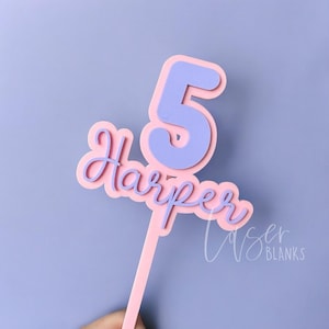 May include: A pink and lilac cake topper featuring the number 5 and the name "Harper" in a cursive font. The number 5 has a lilac centre and a pink outline. The name "Harper" is also pink. The topper is on a pink stick.