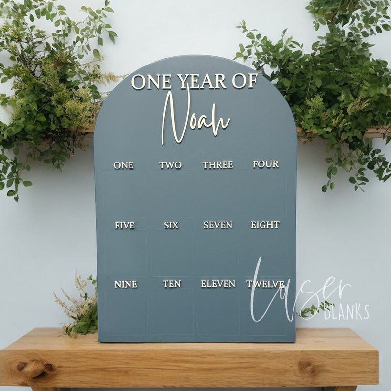 May include: A blue wooden arch-shaped sign with the text "ONE YEAR OF Noah" at the top. The sign has twelve numbered squares, each with a white number from one to twelve.