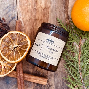 May include: A brown glass jar candle with a label that reads "skön 100% Organic No. 7 Christmas Eve Organic Soy Wax Candle Made with Essential Oils". The candle is surrounded by dried orange slices, cinnamon sticks, and cloves.
