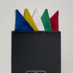 May include: A black pocket square holder with a silver outline of a suit and the text "Fold to Fit". The holder has five pocket squares inside, one each of blue, yellow, white, green, and red.