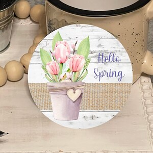 May include: A white wooden tiered stand with a round wooden bead garland, a small potted topiary, a beige mug, and a round coaster with a watercolor illustration of pink tulips in a pot with the text "Hello Spring".