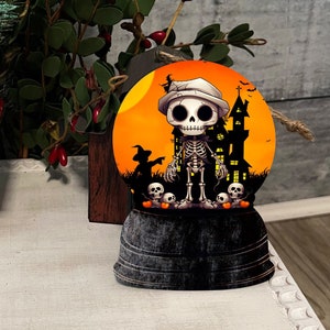 May include: A Halloween-themed snow globe featuring a skeleton wearing a hat, standing in front of a haunted house under an orange moon. The globe sits on a black base, with skulls at the bottom. A red lantern and greenery are in the background.