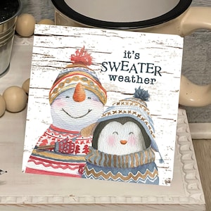 May include: White tiered stand with a snowman and penguin illustration that says "it's sweater weather". The stand has a small potted plant and a wooden bead garland.