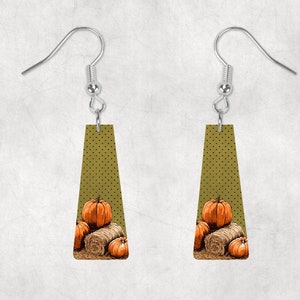 May include: A pair of dangle earrings with a green background and a white polka dot pattern. The earrings feature a design of three orange pumpkins and a bale of hay.