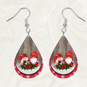 May include: A pair of dangle earrings featuring a red and white winter scene with two gnomes holding a lantern. The earrings are made of wood and have a teardrop shape.
