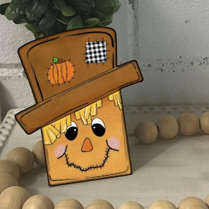 May include: A wooden cutout of a scarecrow with a brown hat, a black and white checkered patch, and a smiling face. The scarecrow is holding a small orange pumpkin. The scarecrow is sitting on a white surface with a wooden bead garland.