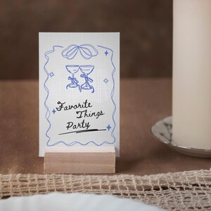 Op de afbeelding: Een witte kaart met blauwe en zwarte tekst en een blauwe illustratie van twee wijnglazen. De kaart staat op een houten standaard en zegt "Favorite Things Party".