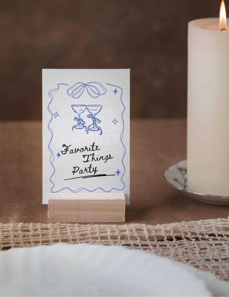 Op de afbeelding: Een witte kaart met blauwe en zwarte tekst en een blauwe illustratie van twee wijnglazen. De kaart staat op een houten standaard en zegt "Favorite Things Party".