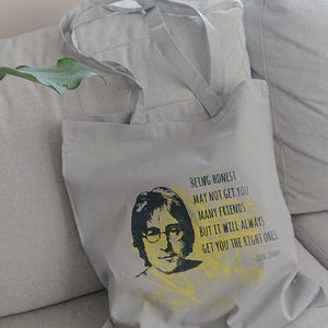Op de afbeelding: Een grijze tote bag met een zwart-wit afbeelding van John Lennon en een citaat dat luidt: "Eerlijk zijn levert je misschien niet veel vrienden op, maar het zal je altijd de juiste mensen opleveren." - John Lennon