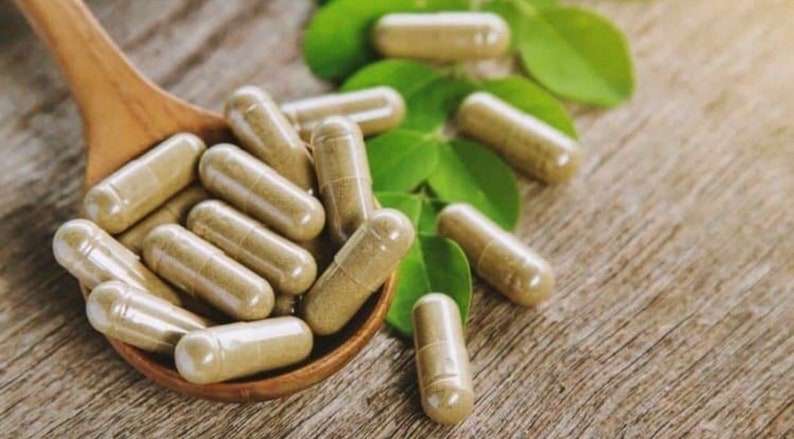 May include: A wooden spoon filled with brown capsules, surrounded by green leaves on a wooden surface.