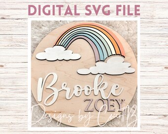 Könnte beinhalten: Ein rundes Holzschild mit einem Regenbogen, Wolken und den Namen "Brooke" und "ZOEY" in weißer Schrift. Das Schild hat einen hellen Holzhintergrund und der Regenbogen besteht aus Pastellfarben.