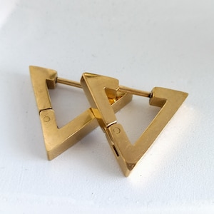 Puede incluir: Un par de pendientes de aro triangulares de oro. Los pendientes están hechos de metal y tienen un diseño simple y geométrico.