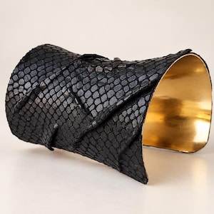 Op de afbeelding: Een brede armband met een zwarte, getextureerde slangenleren buitenkant en een gelaagd, overlappend ontwerp. De binnenkant van de armband is afgewerkt met een gladde, gepolijste goudkleurige metalen voering die contrasteert met het oppervlak.