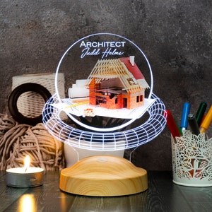 Puede incluir: Un modelo impreso en 3D de una casa en construcción se muestra en una base de luz LED blanca. La base es de madera y tiene un diseño circular. La base de luz está sobre una superficie de madera oscura. El texto "ARCHITECT Judd Helms" es visible en la base de luz.