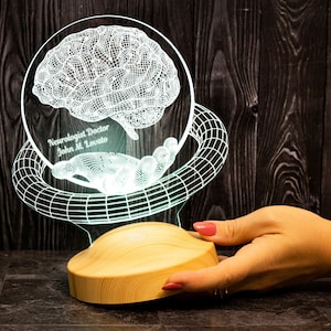 Puede incluir: Un cerebro blanco impreso en 3D y brillante con una base de alambre negro. El cerebro está iluminado desde adentro, creando un brillo blanco suave. La base está hecha de un material acrílico transparente y tiene un diseño de alambre negro. La base está inscrita con el texto "Neurologist Doctor John M. Lovato".