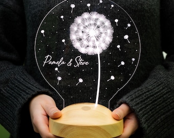May include: A white dandelion with seeds blowing in the wind, illuminated by a light bulb shaped night light. The night light has a wooden base and is personalised with the names "Pamela & Steve".