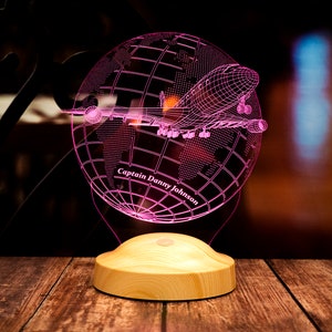 Op de afbeelding: Een roze LED-nachtlamp met een wereldbol en vliegtuigontwerp. De wereldbol is in draadstijl, met een vliegtuig eroverheen. De basis is van lichtgekleurd hout en de tekst "Captain Danny Johnson" is zichtbaar.