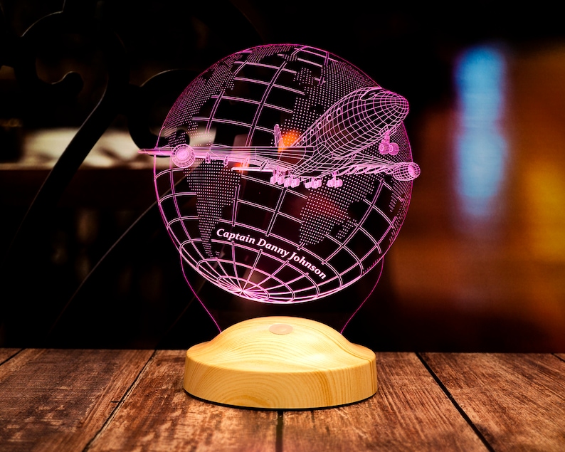 Op de afbeelding: Een roze LED-nachtlamp met een wereldbol en vliegtuigontwerp. De wereldbol is in draadstijl, met een vliegtuig eroverheen. De basis is van lichtgekleurd hout en de tekst "Captain Danny Johnson" is zichtbaar.