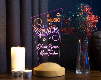 Può includere: Un cartello al neon rosa e arancione con note musicali e il testo "MUSIC" e "Olivia Brown Music Teacher" su una base di legno.