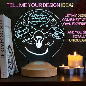 Op de afbeelding: Een lamp die oplicht met een houten voet, met een gloeilamp met een glimlachend gezicht en een gedachteballon die zegt "Laat mijn ontwerper het combineren met zijn eigen ervaring..." en "Vertel me je ontwerpidee?" De lamp staat op een plank met boeken en kaarsen.