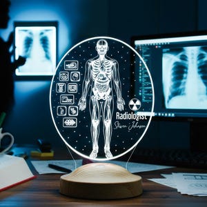 May include: A round, illuminated lamp featuring a human skeletal system illustration. The lamp has a wooden base and includes the text "Radiologist Steven Johnson". The background shows a computer monitor and an X-ray image.