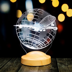 Op de afbeelding: Decoratieve nachtlamp met een wereldbol en een vliegtuigontwerp. De wereldbol is ge&euml;tst met details van de wereldkaart en een vliegtuigsilhouet. De basis is gemaakt van lichtgekleurd hout. De tekst "Captain Danny Johnson" is zichtbaar.