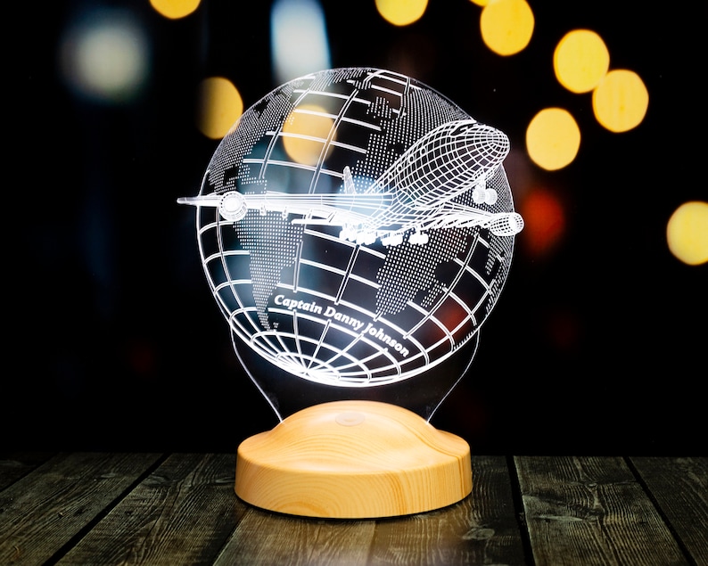 Op de afbeelding: Decoratieve nachtlamp met een wereldbol en een vliegtuigontwerp. De wereldbol is ge&euml;tst met details van de wereldkaart en een vliegtuigsilhouet. De basis is gemaakt van lichtgekleurd hout. De tekst "Captain Danny Johnson" is zichtbaar.