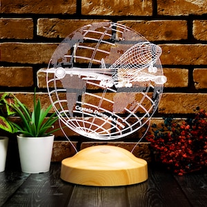 Op de afbeelding: Decoratieve LED-lamp met een wereldbolontwerp en een vliegtuigsilhouet. De lamp is gemaakt van helder acryl en staat op een houten voet. De tekst "Captain Danny Johnson" is zichtbaar op de wereldbol.