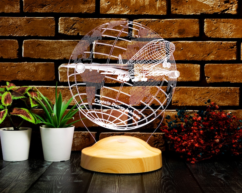 Op de afbeelding: Decoratieve LED-lamp met een wereldbolontwerp en een vliegtuigsilhouet. De lamp is gemaakt van helder acryl en staat op een houten voet. De tekst "Captain Danny Johnson" is zichtbaar op de wereldbol.