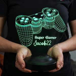 以下が含まれることがあります： 緑色の3Dプリントされたゲームコントローラーランプで、ベースに「Super Gamer Jacob22」の文字が書かれています。