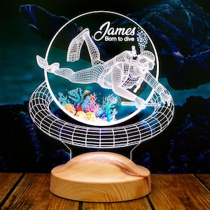 Könnte beinhalten: Ein weißes, 3D-gedrucktes Nachtlicht in Form eines Tauchers mit Korallenriff und Fischen. Die Lampe hat einen Holzsockel und der Text "James Born to dive" ist auf die Lampe gedruckt.
