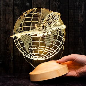 Op de afbeelding: Een verlichte nachtlamp met een vliegtuig dat rond een wereldbol vliegt. De lamp is gemaakt van helder acryl en heeft een houten voet. De tekst "Captain Danny Johnson" is zichtbaar op de wereldbol.