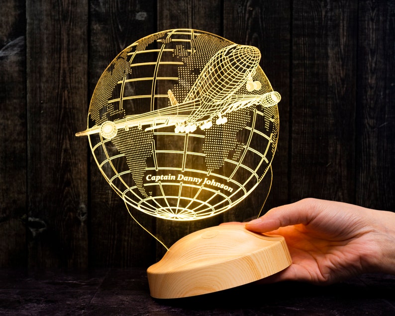 Op de afbeelding: Een verlichte nachtlamp met een vliegtuig dat rond een wereldbol vliegt. De lamp is gemaakt van helder acryl en heeft een houten voet. De tekst "Captain Danny Johnson" is zichtbaar op de wereldbol.