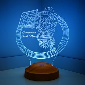 May include: A blue 3D printed lamp shaped like a film camera with the text "Cameraman Sarah Moon" on the base. The lamp is on a wooden base.