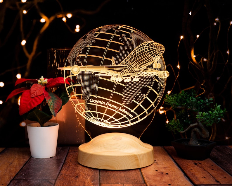 Op de afbeelding: Een gloeiende nachtlamp in de vorm van een wereldbol met een vliegtuigontwerp. De tekst "Captain Danny Johnson" is gegraveerd. De basis is van hout. De achtergrond is donker, met lichtjes en planten.