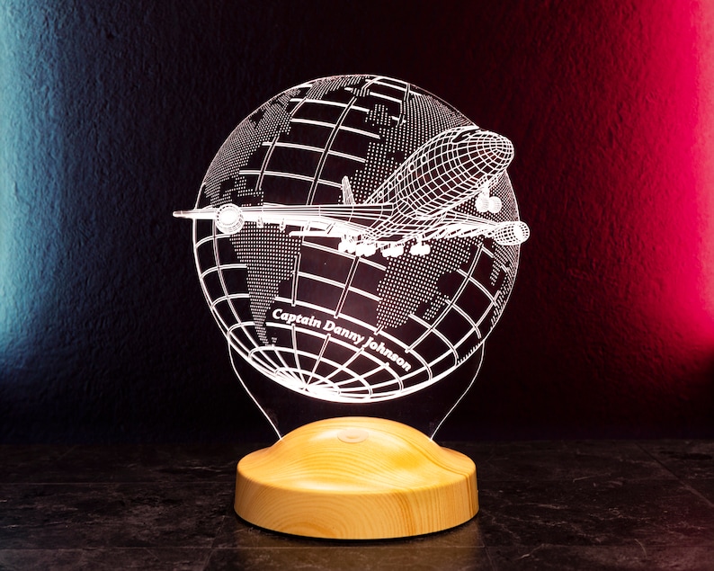 Op de afbeelding: Een decoratieve LED-lamp met een wereldbol en vliegtuigontwerp. De wereldbol is ge&euml;tst met continenten en het vliegtuig is in vlucht. De lamp bevat de tekst "Captain Danny Johnson" en staat op een houten voet. De achtergrond is donker met rode en blauwe verlichting.