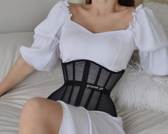 May include: A black mesh corset with boning and a white label that says "@corset_pv" on a woman wearing a white blouse with puffed sleeves.