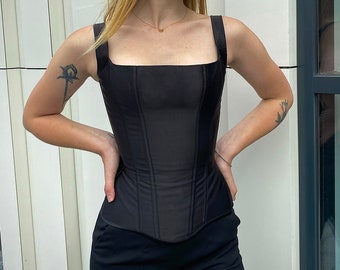 May include: A black corset with a square neckline and boning details. The corset is made of a smooth, satin-like fabric. The model is wearing the corset with black pants.
