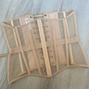 May include: A beige mesh corset with a lace-up back and a satin ribbon detail. The corset has a structured design with multiple panels and a curved bottom.