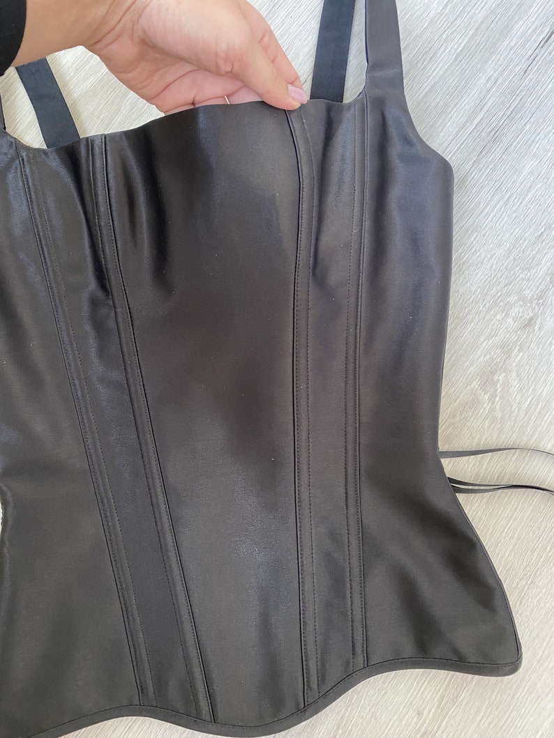 May include: A black satin corset with boning and adjustable straps. The corset has a low neckline and a pointed bottom.