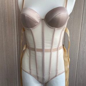 May include: A beige bodysuit with a structured bustier top and sheer mesh panels. The garment features adjustable straps and gold-colored ribbon ties. The bustier has a satin finish, and the bodysuit is displayed on a mannequin.