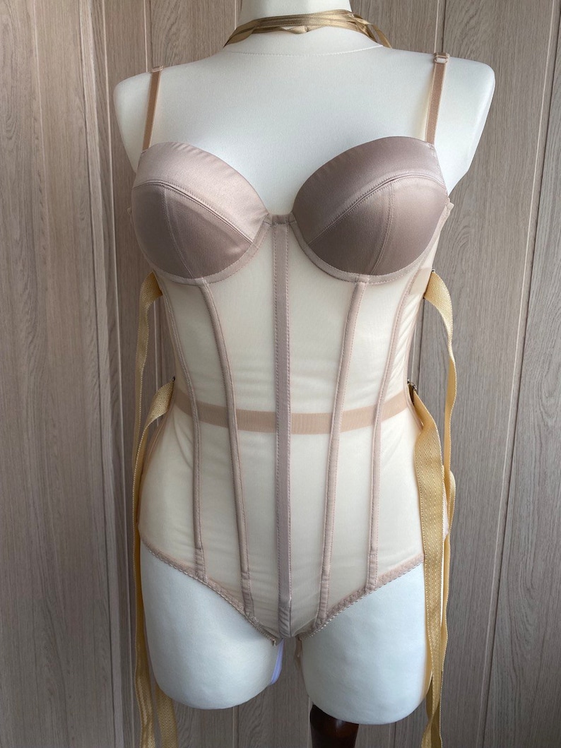 May include: A beige bodysuit with a structured bustier top and sheer mesh panels. The garment features adjustable straps and gold-colored ribbon ties. The bustier has a satin finish, and the bodysuit is displayed on a mannequin.