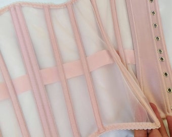 May include: A pink corset with a mesh overlay and a row of metal eyelets on the side. The corset has a lace trim and is made of a soft, stretchy fabric.