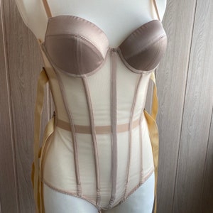 May include: A beige corset bodysuit displayed on a mannequin. The bodice features a structured, padded bust with adjustable straps. The sheer mesh body is accented with vertical boning and gold ribbon ties.