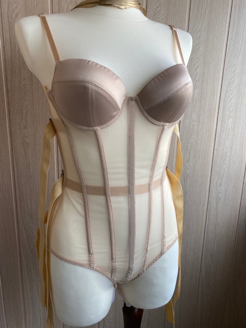 May include: A beige corset bodysuit displayed on a mannequin. The bodice features a structured, padded bust with adjustable straps. The sheer mesh body is accented with vertical boning and gold ribbon ties.
